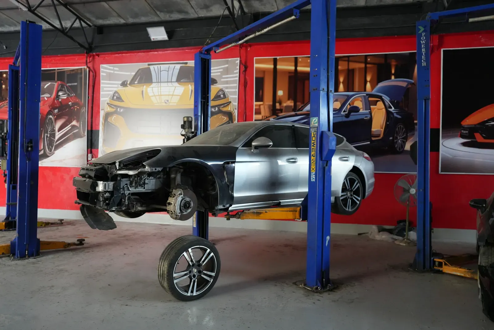 A car raised on a two-post lift in the workshop with the front bumper removed and the front wheel off, exposing the brake disc and hub.
