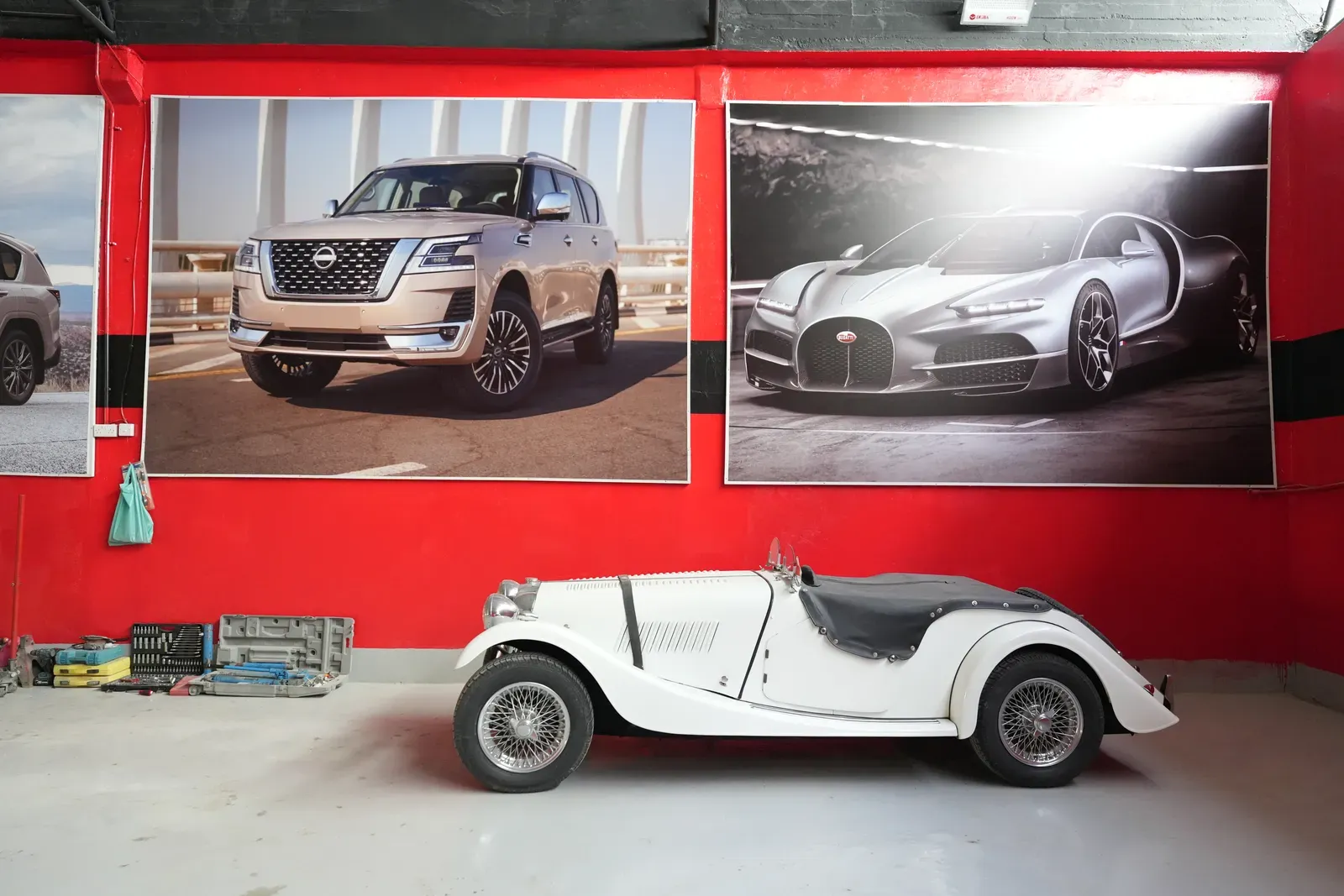 A white classic roadster standing against the red workshop wall in Sharjah, beneath full-height prints of a Nissan Patrol and a Bugatti.