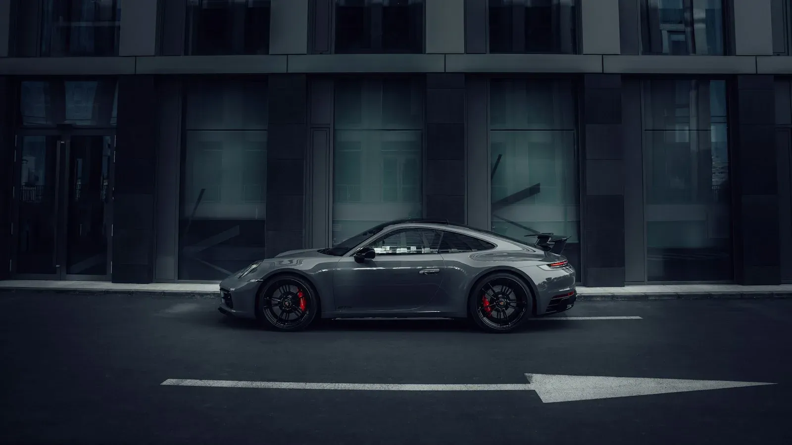 A grey Porsche 911 in side profile against a dark glazed facade.
